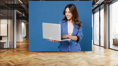 Smiling cheerful funny beautiful attractive young brunette woman 20s wearing basic casual jacket standing working on laptop pc computer isolated on bright blue colour background studio portrait. Wall mural