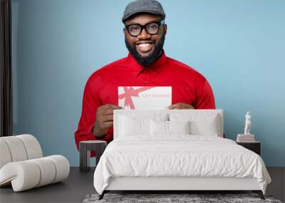 Smiling cheerful funny young bearded african american man 20s wearing casual red shirt eyeglasses cap standing hold gift certificate isolated on pastel blue color wall background studio portrait. Wall mural