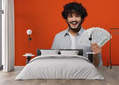 Smiling cheerful happy young bearded Indian man 20s years old wears blue shirt holding showing fan of cash money in dollar banknotes looking camera isolated on plain orange background studio portrait. Wall mural