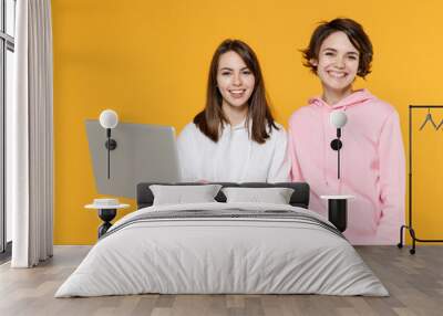 Smiling cheerful two young women friends 20s wearing casual white pink hoodies standing working on laptop pc computer looking camera isolated on bright yellow color wall background studio portrait. Wall mural