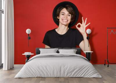 Smiling cheerful young brunette woman 20s years old wearing casual basic black t-shirt hat standing showing ok okay gesture looking camera isolated on bright red color background studio portrait. Wall mural