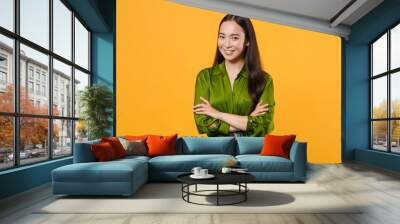 Smiling confident beautiful attractive young brunette asian woman wearing basic green shirt standing holding hands crossed looking camera isolated on bright yellow colour background, studio portrait. Wall mural