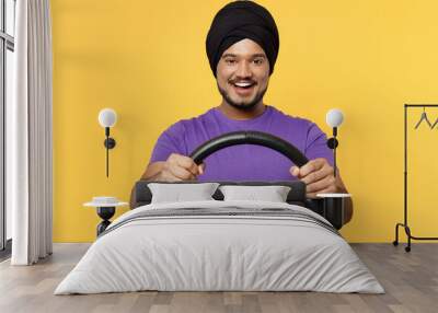 Smiling devotee Sikh Indian man ties his traditional turban dastar wears purple t-shirt hold steering wheel driving car isolated on plain yellow background studio portrait. People lifestyle concept. Wall mural