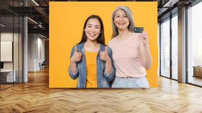 Smiling family asian female women girls gray-haired mother and brunette daughter in casual clothes posing hold credit bank card showing thumbs up isolated on yellow color background studio portrait. Wall mural