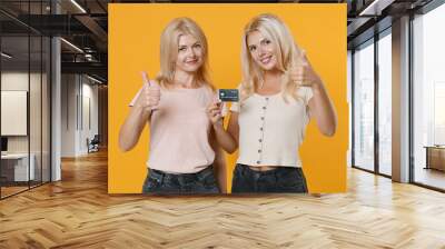 Smiling family two women blonde mother and daughter wearing casual clothes standing holding in hand credit bank card showing thumbs up isolated on bright yellow colour background studio portrait. Wall mural