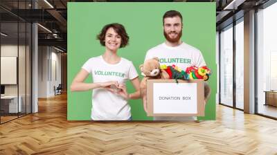 Smiling friends couple in volunteer t-shirt isolated on green background. Voluntary free work assistance help charity grace teamwork. Hold donation box with kids toys, showing shape heart with hands. Wall mural