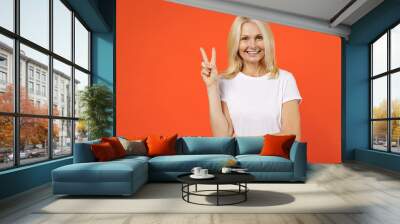 Smiling funny elderly gray-haired blonde woman lady 40s 50s years old in white casual t-shirt standing showing victory sign looking camera isolated on bright orange color background studio portrait. Wall mural