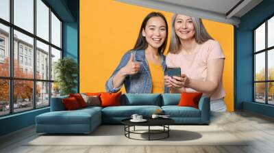 Smiling funny family asian female women gray-haired mother brunette daughter in casual clothes posing using mobile cell phone showing thumb up isolated on yellow color wall background studio portrait. Wall mural