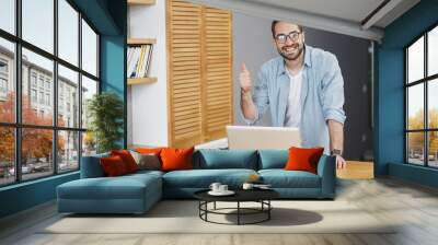 Smiling funny handsome young bearded business man 20s in blue shirt glasses standing near desk with cup of coffee or tea working on laptop pc computer showing thumb up spending time at home or office. Wall mural