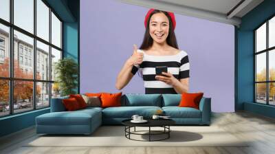 Smiling funny young asian woman 20s wearing striped t-shirt red beret using mobile cell phone typing sms message showing thumb up looking camera isolated on pastel violet background studio portrait. Wall mural