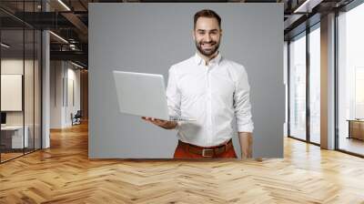 Smiling handsome young bearded business man in classic white shirt working on laptop pc computer looking camera isolated on grey color background studio portrait. Achievement career wealth concept. Wall mural