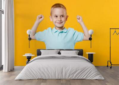 Smiling happy little fun male fair-haired brown-eyed kid boy 5-6 years old wearing stylish blue turquoise t-shirt polo raised hands up isolated on yellow color wall background, child studio portrait. Wall mural