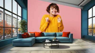 Smiling joyful young brunette woman girl in yellow sweater posing isolated on pastel pink background studio portrait. People sincere emotions lifestyle concept. Mock up copy space. Clenching fists. Wall mural