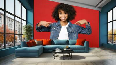 Smiling little african american kid girl 12-13 years old in casual denim jacket pointing index fingers on camera isolated on red color background children studio portrait. Childhood lifestyle concept. Wall mural