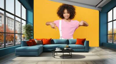 Smiling little african american kid girl 12-13 years old in pink t-shirt isolated on yellow background studio portrait. Childhood lifestyle concept. Mock up copy space. Pointing index fingers down. Wall mural