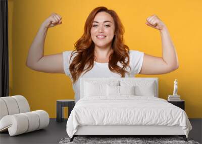 Smiling strong young redhead plus size body positive female woman girl 20s in white casual t-shirt posing showing biceps muscles looking camera isolated on yellow color background studio portrait. Wall mural