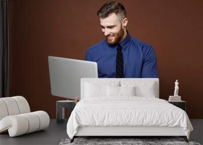 Smiling successful bearded young business man wearing blue shirt tie working on laptop pc computer isolated on brown colour background studio portrait. Achievement career wealth business concept. Wall mural