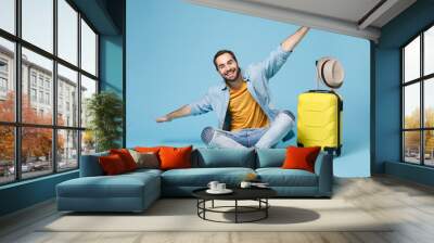 Smiling traveler tourist man in yellow clothes isolated on blue wall background. Male passenger traveling abroad on weekend. Air flight journey concept. Sit near suitcase, spreading hands like flying. Wall mural