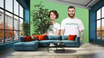 Smiling two young friends couple in white volunteer t-shirt isolated on pastel green background in studio. Voluntary free work assistance help charity grace teamwork concept. Hold green flowerpots. Wall mural