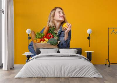 Smiling woman in denim clothes isolated on yellow orange background. Delivery service from shop or restaurant concept. Hold brown craft paper bag for takeaway mock up with food products, bell pepper. Wall mural