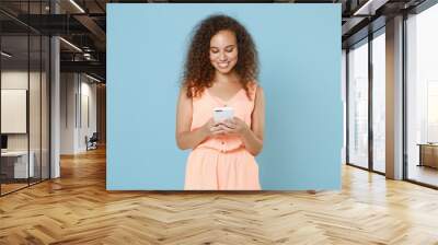 Smiling young african american girl in pastel summer clothes isolated on blue wall background studio portrait. People lifestyle concept. Mock up copy space. Using mobile cell phone typing sms message. Wall mural