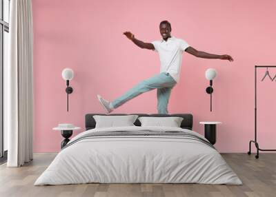 Smiling young african american man guy in white polo shirt, turquoise trousers isolated on pastel pink background. People lifestyle concept. Mock up copy space. Spreading hands, rising leg, dancing. Wall mural