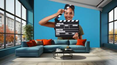 Smiling young african american woman girl in casual t-shirt posing isolated on bright blue background. People lifestyle concept. Mock up copy space. Holding classic black film making clapperboard. Wall mural