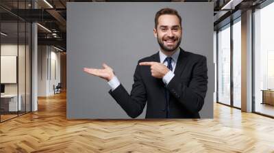 Smiling young bearded business man in classic black suit shirt tie posing isolated on grey background. Achievement career wealth business concept. Mock up copy space. Pointing index finger hand aside. Wall mural