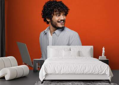 Smiling young bearded Indian man 20s years old wear blue shirt hold use work on laptop pc computer typing browsing chatting send sms looking behind isolated on plain orange background studio portrait Wall mural