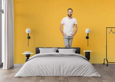 Smiling young bearded man guy in white casual t-shirt posing isolated on yellow background studio portrait. People sincere emotions lifestyle concept. Mock up copy space. Holding hands in pockets. Wall mural