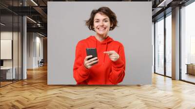 Smiling young brunette woman girl in casual red hoodie posing isolated on grey wall background studio portrait. People lifestyle concept. Mock up copy space. Pointing index finger on mobile phone. Wall mural