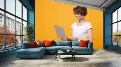 Smiling young brunette woman girl in white t-shirt posing isolated on yellow orange background studio portrait. People emotions lifestyle concept. Mock up copy space. Working on laptop pc computer. Wall mural