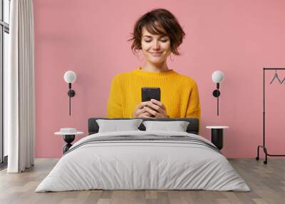 Smiling young brunette woman girl in yellow sweater posing isolated on pastel pink wall background studio portait. People lifestyle concept. Mock up copy space. Using mobile phone, typing sms message. Wall mural