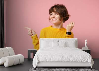 Smiling young brunette woman girl in yellow sweater posing isolated on pink background in studio. People lifestyle concept. Mock up copy space. Wearing smart watch on hand pointing index finger aside. Wall mural