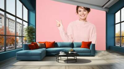 Smiling young brunette woman girl wearing knitted casual sweater posing isolated on pastel pink background studio portrait. People lifestyle concept. Mock up copy space. Pointing index finger aside. Wall mural