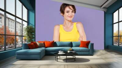 Smiling young brunette woman girl wearing yellow casual tank top posing isolated on pastel violet wall background studio portrait. People sincere emotions lifestyle concept. Holding hands crossed. Wall mural