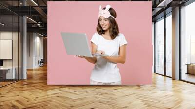 Smiling young brunette woman in white pajamas home wear sleep mask working on laptop pc computer resting at home isolated on pastel pink background studio portrait. Relax good mood lifestyle concept. Wall mural