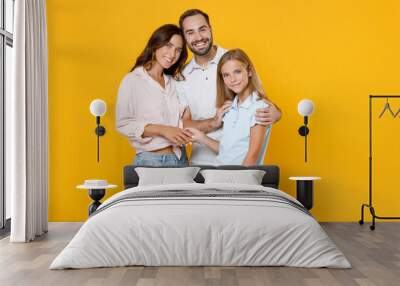 Smiling young happy parents mom dad with child kid daughter teen girl in basic t-shirts hugging looking camera isolated on yellow background studio portrait. Family day parenthood childhood concept. Wall mural