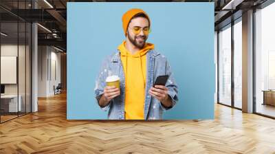 Smiling young hipster guy in fashion jeans denim clothes posing isolated on pastel blue background. People lifestyle concept. Mock up copy space. Using mobile phone, hold paper cup of coffee or tea. Wall mural