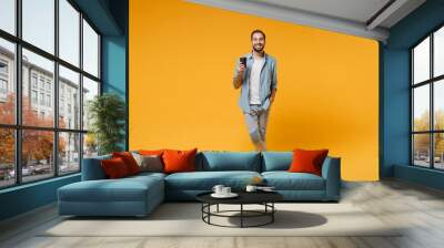 Smiling young man in casual blue shirt posing isolated on yellow orange wall background studio portrait. People sincere emotions lifestyle concept. Mock up copy space. Hold paper cup of coffee or tea. Wall mural