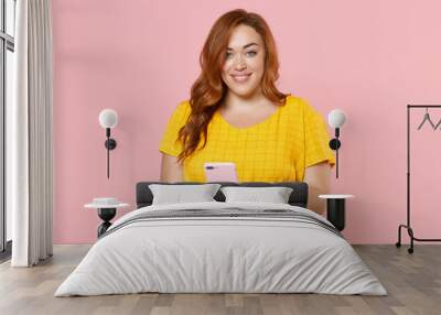 Smiling young redhead plus size body positive female woman girl 20s in yellow dress posing using mobile cell phone typing sms message isolated on pastel pink color wall background studio portrait. Wall mural