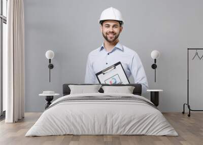 Smiling young unshaven business man in light shirt, protective construction white helmet isolated on grey background. Achievement career wealth business concept. Hold clipboard with papers document. Wall mural