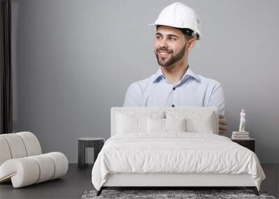 Smiling young unshaven business man in light shirt protective construction helmet isolated on grey wall background. Achievement career wealth business concept. Holding hands crossed, looking aside. Wall mural