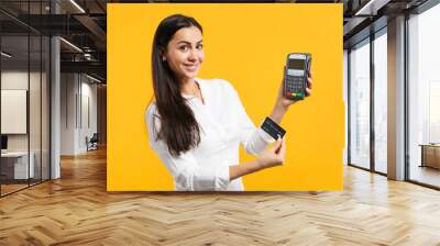 Smiling young woman holding wireless modern bank payment terminal to process and acquire credit card payments isolated on yellow orange wall background. People lifestyle concept. Mock up copy space. Wall mural