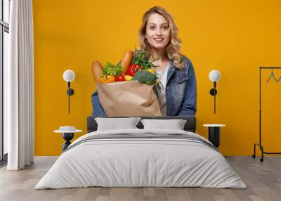 Smiling young woman in denim clothes posing isolated on yellow orange background. Delivery service from shop or restaurant concept. Hold brown craft paper bag for takeaway mock up with food products. Wall mural