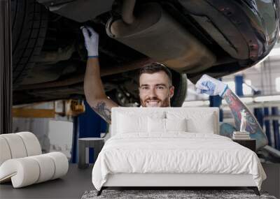 Strong young male professional technician mechanic man wears denim blue overalls t-shirt show muscles strength stand near car lift check technical condition work in vehicle repair shop workshop inside Wall mural