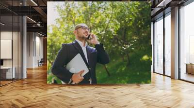 Successful smart businessman in white shirt, classic suit, glasses. Man stand with laptop pc computer, talk on mobile phone in city park outdoors on nature background. Mobile Office, business concept. Wall mural