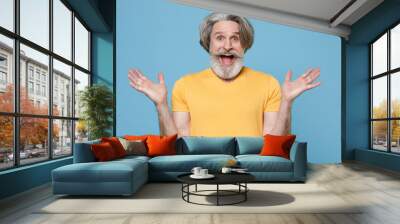 Surprised elderly gray-haired mustache bearded man in casual yellow t-shirt posing isolated on blue background studio. People lifestyle concept. Mock up copy space. Keeping mouth open spreading hands. Wall mural