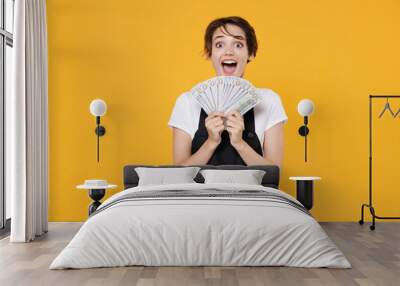 Surprised excited young female woman 20s barista bartender barman employee in t-shirt apron posing holding in hands fan of cash money in dollar banknotes isolated on yellow background studio portrait. Wall mural