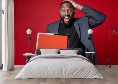 Surprised young african american business man 20s in classic jacket suit standing put hand on head hold laptop pc computer with blank empty screen isolated on red color background studio portrait. Wall mural